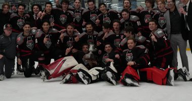 University of New Mexico Lobos in black and red uniforms poses on the ice with a championship trophy, celebrating at The Cube rink after a playoff win