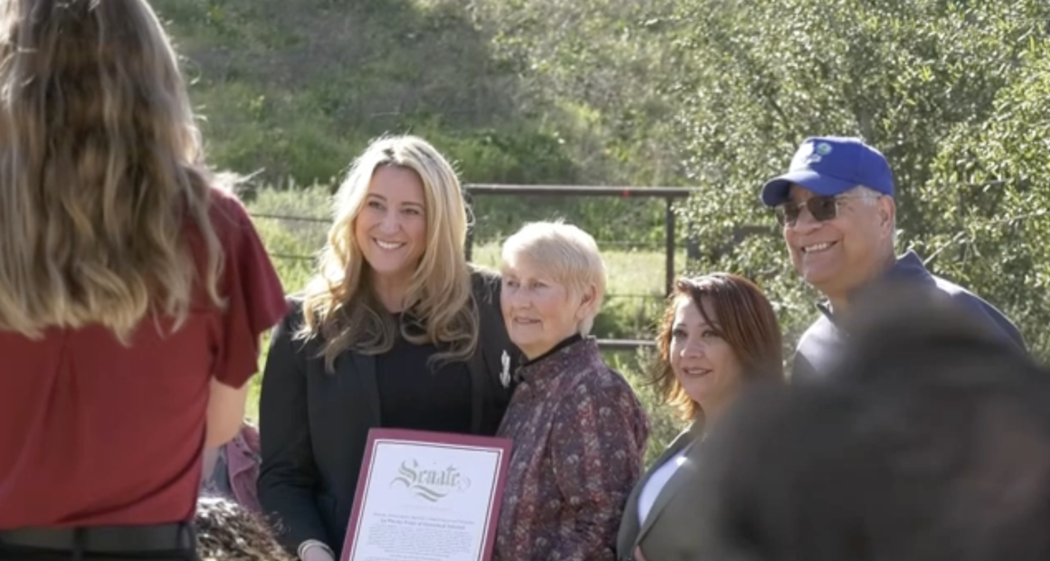 **Alt text:** A group of four people stand outdoors in a sunny, grassy area while posing for a photo. A woman in a black blazer holds a framed certificate that reads “Senate.” Beside her are an older woman with short gray hair, another woman with reddish-brown hair, and a man wearing a blue baseball cap and sunglasses. A photographer stands in the foreground taking the picture, while trees and a fence appear in the background.