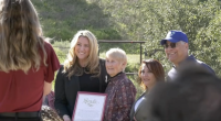 **Alt text:** A group of four people stand outdoors in a sunny, grassy area while posing for a photo. A woman in a black blazer holds a framed certificate that reads “Senate.” Beside her are an older woman with short gray hair, another woman with reddish-brown hair, and a man wearing a blue baseball cap and sunglasses. A photographer stands in the foreground taking the picture, while trees and a fence appear in the background.