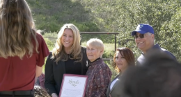 **Alt text:** A group of four people stand outdoors in a sunny, grassy area while posing for a photo. A woman in a black blazer holds a framed certificate that reads “Senate.” Beside her are an older woman with short gray hair, another woman with reddish-brown hair, and a man wearing a blue baseball cap and sunglasses. A photographer stands in the foreground taking the picture, while trees and a fence appear in the background.