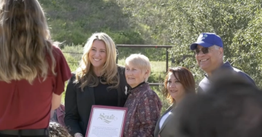 **Alt text:** A group of four people stand outdoors in a sunny, grassy area while posing for a photo. A woman in a black blazer holds a framed certificate that reads “Senate.” Beside her are an older woman with short gray hair, another woman with reddish-brown hair, and a man wearing a blue baseball cap and sunglasses. A photographer stands in the foreground taking the picture, while trees and a fence appear in the background.