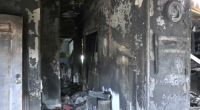 Interior hallway of a fire-damaged home with charred black walls, peeling paint, and debris scattered across the floor, with daylight visible through an opening at the far end.