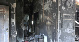 Interior hallway of a fire-damaged home with charred black walls, peeling paint, and debris scattered across the floor, with daylight visible through an opening at the far end.