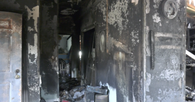 Interior hallway of a fire-damaged home with charred black walls, peeling paint, and debris scattered across the floor, with daylight visible through an opening at the far end.