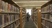 Alt text: Person standing in a library aisle reaching for a book on a shelf, with tall bookshelves filled with books on both sides and a study area visible near a bright window at the end of the aisle.