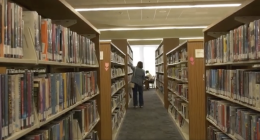 Alt text: Person standing in a library aisle reaching for a book on a shelf, with tall bookshelves filled with books on both sides and a study area visible near a bright window at the end of the aisle.