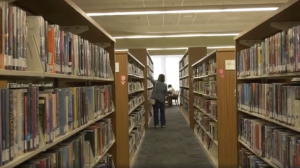 Alt text: Person standing in a library aisle reaching for a book on a shelf, with tall bookshelves filled with books on both sides and a study area visible near a bright window at the end of the aisle.
