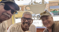 Danny Ramirez(center) Roy Ruiz(left) and a third man on the right pose for a picture with iced coffee in a beige colored coffee shop