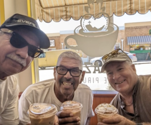 Danny Ramirez(center) Roy Ruiz(left) and a third man on the right pose for a picture with iced coffee in a beige colored coffee shop