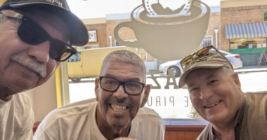 Danny Ramirez(center) Roy Ruiz(left) and a third man on the right pose for a picture with iced coffee in a beige colored coffee shop