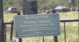 Sign at the entrance to Ed Davis Park at Towsley Canyon in Santa Clarita, identifying it as part of the Santa Clarita Woodlands Park system, with cars and grassy hills visible in the background.
