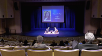 Audience members sit in a large auditorium watching a stage discussion, where two speakers are seated on chairs under blue lighting. A large screen behind them displays a portrait and the name “Dr. Jasmine Ruys” along with event branding.