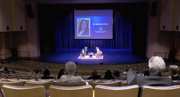Audience members sit in a large auditorium watching a stage discussion, where two speakers are seated on chairs under blue lighting. A large screen behind them displays a portrait and the name “Dr. Jasmine Ruys” along with event branding.
