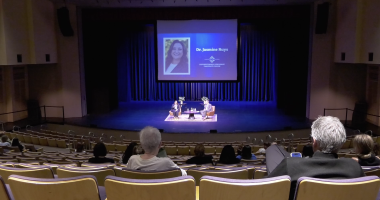 Audience members sit in a large auditorium watching a stage discussion, where two speakers are seated on chairs under blue lighting. A large screen behind them displays a portrait and the name “Dr. Jasmine Ruys” along with event branding.