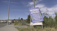 A roadside sign reading “Entrance – Haskell Canyon Bike Park Project” stands tilted on a patch of grass beside a sidewalk. The sign indicates access for contractors and authorized personnel only, with a suburban street, houses, and utility poles visible in the background under a blue sky.