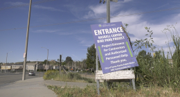 A roadside sign reading “Entrance – Haskell Canyon Bike Park Project” stands tilted on a patch of grass beside a sidewalk. The sign indicates access for contractors and authorized personnel only, with a suburban street, houses, and utility poles visible in the background under a blue sky.