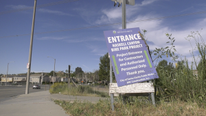 A roadside sign reading “Entrance – Haskell Canyon Bike Park Project” stands tilted on a patch of grass beside a sidewalk. The sign indicates access for contractors and authorized personnel only, with a suburban street, houses, and utility poles visible in the background under a blue sky.