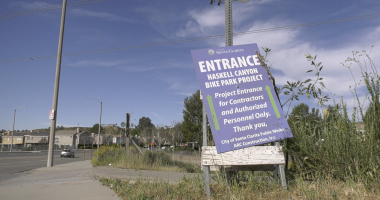 A roadside sign reading “Entrance – Haskell Canyon Bike Park Project” stands tilted on a patch of grass beside a sidewalk. The sign indicates access for contractors and authorized personnel only, with a suburban street, houses, and utility poles visible in the background under a blue sky.