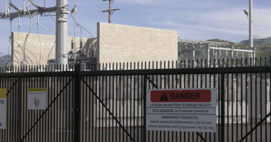 A fenced industrial facility with electrical equipment and power lines, featuring a prominent warning sign that reads “Danger: Lithium Ion Battery Storage Facility,” along with an address and emergency contact numbers. Hills and a partly cloudy sky are visible in the background.