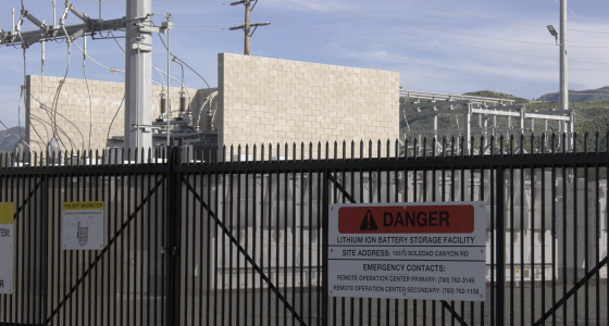 A fenced industrial facility with electrical equipment and power lines, featuring a prominent warning sign that reads “Danger: Lithium Ion Battery Storage Facility,” along with an address and emergency contact numbers. Hills and a partly cloudy sky are visible in the background.