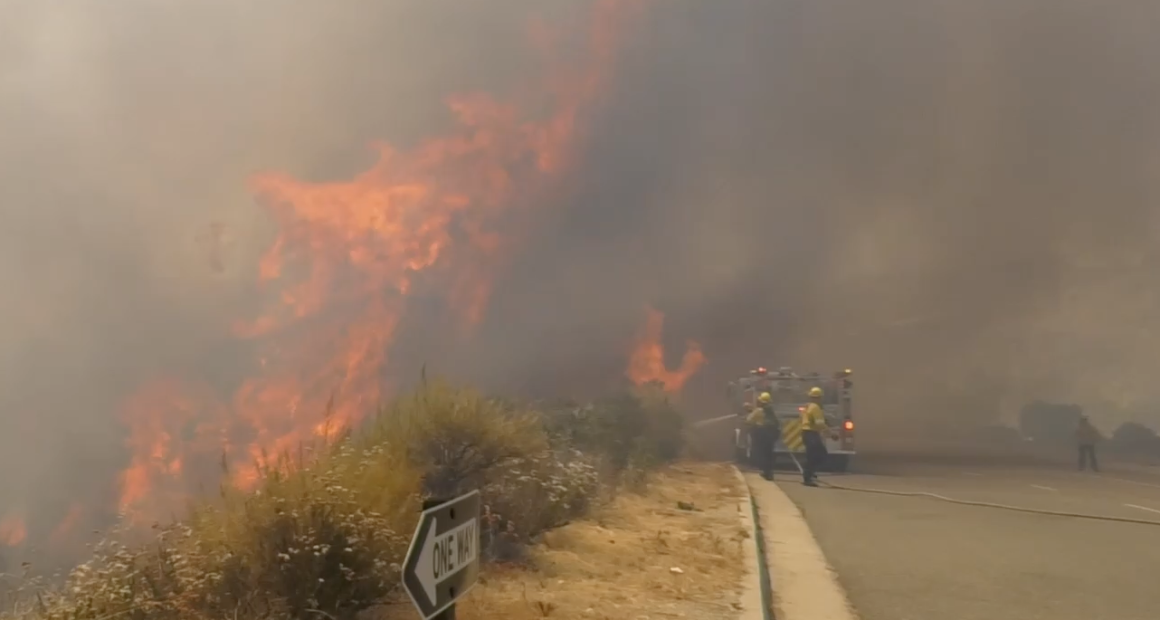Firefighters in yellow gear battle a large roadside wildfire as tall flames and thick smoke engulf dry brush next to a paved road, with a fire engine parked nearby and visibility reduced by haze.