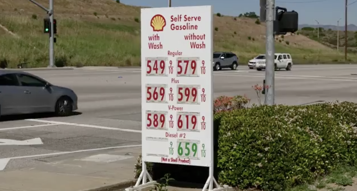 A roadside Shell gas station sign displays high fuel prices—regular, plus, V-Power, and diesel—at an intersection, with cars passing by and dry hills in the background.