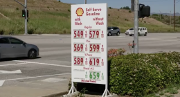 A roadside Shell gas station sign displays high fuel prices—regular, plus, V-Power, and diesel—at an intersection, with cars passing by and dry hills in the background.