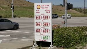 A roadside Shell gas station sign displays high fuel prices—regular, plus, V-Power, and diesel—at an intersection, with cars passing by and dry hills in the background.
