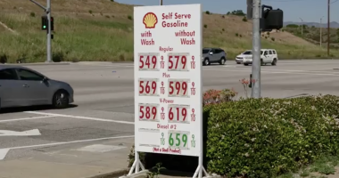 A roadside Shell gas station sign displays high fuel prices—regular, plus, V-Power, and diesel—at an intersection, with cars passing by and dry hills in the background.