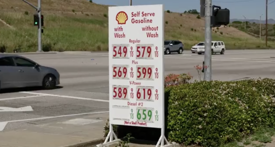 A roadside Shell gas station sign displays high fuel prices—regular, plus, V-Power, and diesel—at an intersection, with cars passing by and dry hills in the background.
