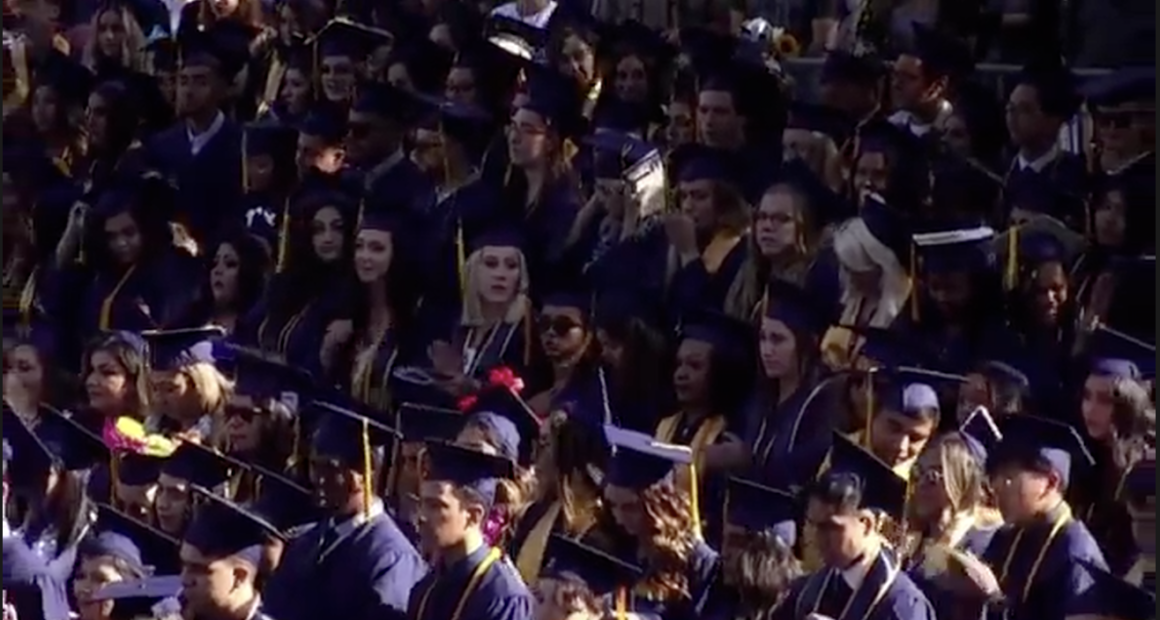 A large group of graduates in caps and gowns stand closely together during an outdoor commencement ceremony, many wearing honor cords and looking toward the stage.