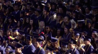 A large group of graduates in caps and gowns stand closely together during an outdoor commencement ceremony, many wearing honor cords and looking toward the stage.