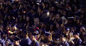 A large group of graduates in caps and gowns stand closely together during an outdoor commencement ceremony, many wearing honor cords and looking toward the stage.