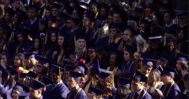 A large group of graduates in caps and gowns stand closely together during an outdoor commencement ceremony, many wearing honor cords and looking toward the stage.