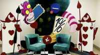 Colorful Alice in Wonderland-themed display featuring the Cheshire Cat grinning behind a large teacup and a tilted “10/6” hat sign, flanked by two oversized playing card guards holding spears. Two teal velvet chairs sit in front on a black-and-white checkered floor, creating a whimsical photo backdrop.