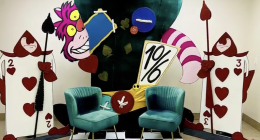 Colorful Alice in Wonderland-themed display featuring the Cheshire Cat grinning behind a large teacup and a tilted “10/6” hat sign, flanked by two oversized playing card guards holding spears. Two teal velvet chairs sit in front on a black-and-white checkered floor, creating a whimsical photo backdrop.