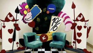 Colorful Alice in Wonderland-themed display featuring the Cheshire Cat grinning behind a large teacup and a tilted “10/6” hat sign, flanked by two oversized playing card guards holding spears. Two teal velvet chairs sit in front on a black-and-white checkered floor, creating a whimsical photo backdrop.