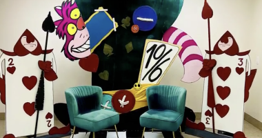 Colorful Alice in Wonderland-themed display featuring the Cheshire Cat grinning behind a large teacup and a tilted “10/6” hat sign, flanked by two oversized playing card guards holding spears. Two teal velvet chairs sit in front on a black-and-white checkered floor, creating a whimsical photo backdrop.