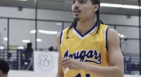 Donovan Chapman wearing a yellow “Cougars” jersey with the number 14 stands on an indoor court, looking focused during gameplay. He has braided hair and facial hair, with gym bleachers and other players visible blurred in the background.