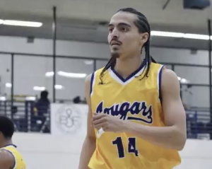 Donovan Chapman wearing a yellow “Cougars” jersey with the number 14 stands on an indoor court, looking focused during gameplay. He has braided hair and facial hair, with gym bleachers and other players visible blurred in the background.