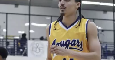 Donovan Chapman wearing a yellow “Cougars” jersey with the number 14 stands on an indoor court, looking focused during gameplay. He has braided hair and facial hair, with gym bleachers and other players visible blurred in the background.