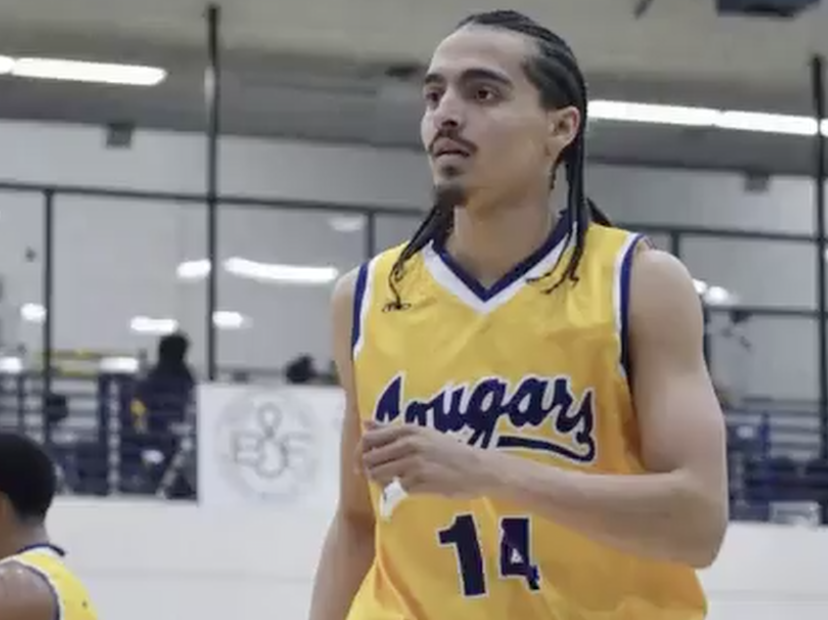 Donovan Chapman wearing a yellow “Cougars” jersey with the number 14 stands on an indoor court, looking focused during gameplay. He has braided hair and facial hair, with gym bleachers and other players visible blurred in the background.