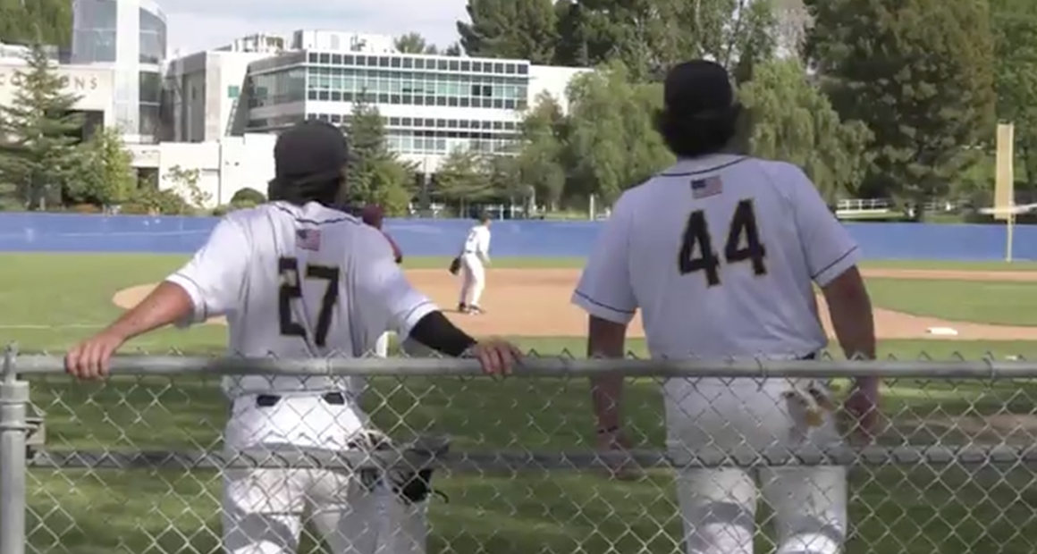 Two College of the Canyons baseball players in white uniforms, numbers 27 and 44, lean on a chain-link fence watching the game on the field, with a pitcher visible in the distance and campus buildings and trees in the background.