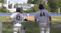 Two College of the Canyons baseball players in white uniforms, numbers 27 and 44, lean on a chain-link fence watching the game on the field, with a pitcher visible in the distance and campus buildings and trees in the background.