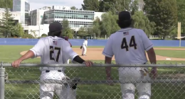 Two College of the Canyons baseball players in white uniforms, numbers 27 and 44, lean on a chain-link fence watching the game on the field, with a pitcher visible in the distance and campus buildings and trees in the background.