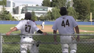 Two College of the Canyons baseball players in white uniforms, numbers 27 and 44, lean on a chain-link fence watching the game on the field, with a pitcher visible in the distance and campus buildings and trees in the background.