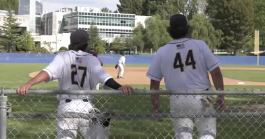 Two College of the Canyons baseball players in white uniforms, numbers 27 and 44, lean on a chain-link fence watching the game on the field, with a pitcher visible in the distance and campus buildings and trees in the background.