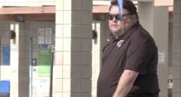 A Sean Kakumu sunglasses and a dark polo shirt stands outdoors near tiled columns and a railing, with a blue object partially obscuring the foreground.