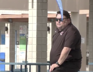 A Sean Kakumu sunglasses and a dark polo shirt stands outdoors near tiled columns and a railing, with a blue object partially obscuring the foreground.