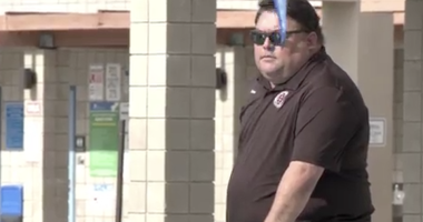 A Sean Kakumu sunglasses and a dark polo shirt stands outdoors near tiled columns and a railing, with a blue object partially obscuring the foreground.